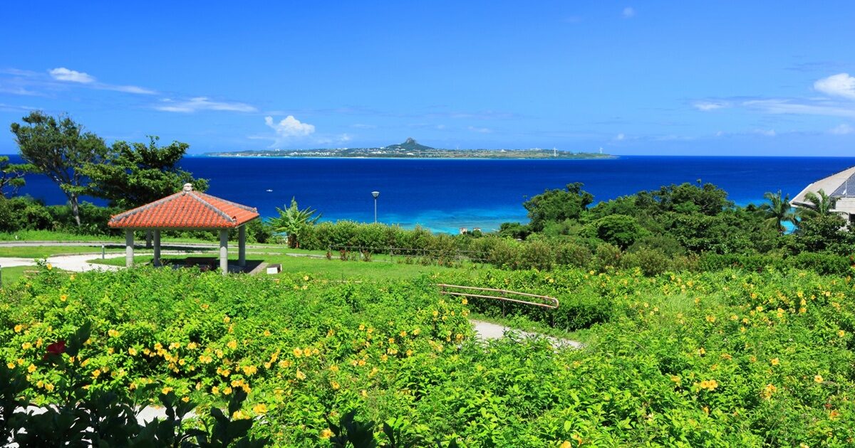黄色い花咲く庭園と赤瓦屋根のあずまや、遠くに見える伊江島